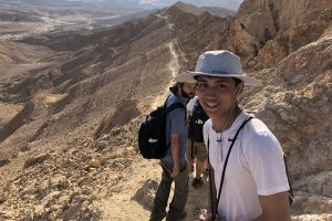 Some Bronfman Fellows are on a path in the desert in Israel, with mountains in the background. One looks back and smiles at the camera.