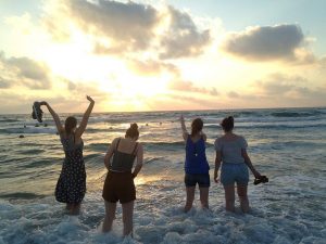 Four Fellows on the beach in Israel, with their feet in the water. They are silhouetted against the sun. One of them holds up her hands.