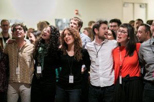 A group of Bronfman alumni stand with their arms around each other and sing together.