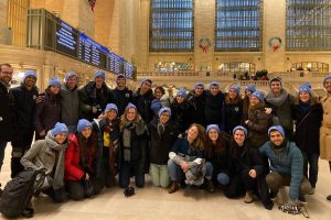 A group of Israeli Bronfman Fellows pose in Grand Central Station. They are dressed in winter clothes and all of them wear blue hats.