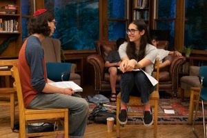 Two Fellows, a male and a female, sit and talk together with books on their laps.  The girl smiles at the boy, who wears a kippah.