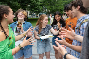 A group of young people, male and female, are standing in a park. They are laughing and pointing excitedly at each other.